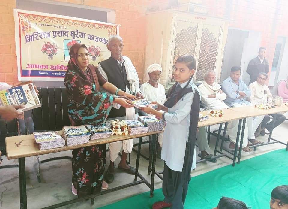 A student in school uniform receiving books across a table stacked with more books, under a Giriraj Prasad Ghuraiya Foundation banner.