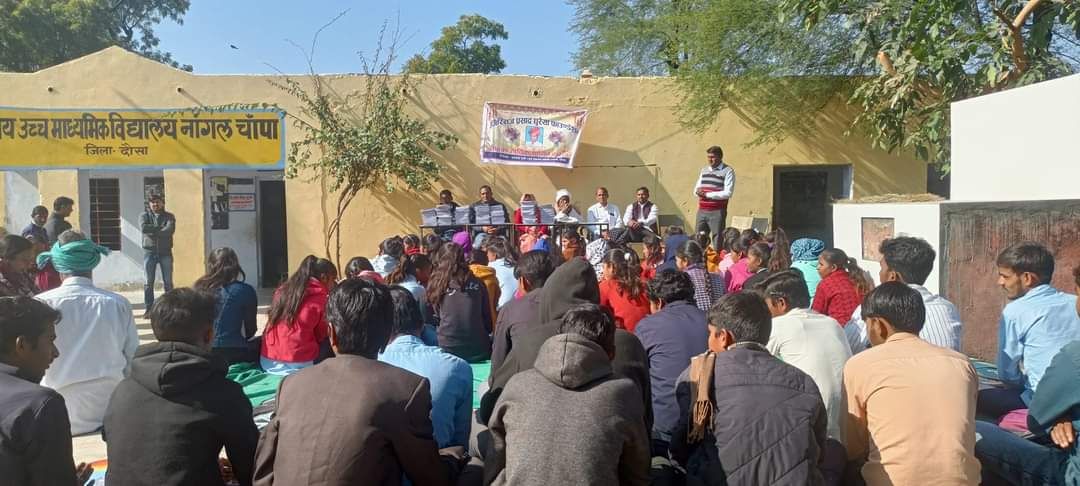 Students and villagers seated on mats in a school courtyard facing a row of speakers, with a banner strung across the wall behind them.