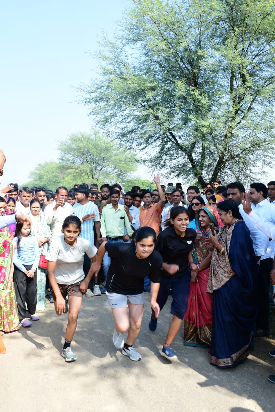 Three young women running on a village road at the start of a race, with a large crowd of spectators on both sides.