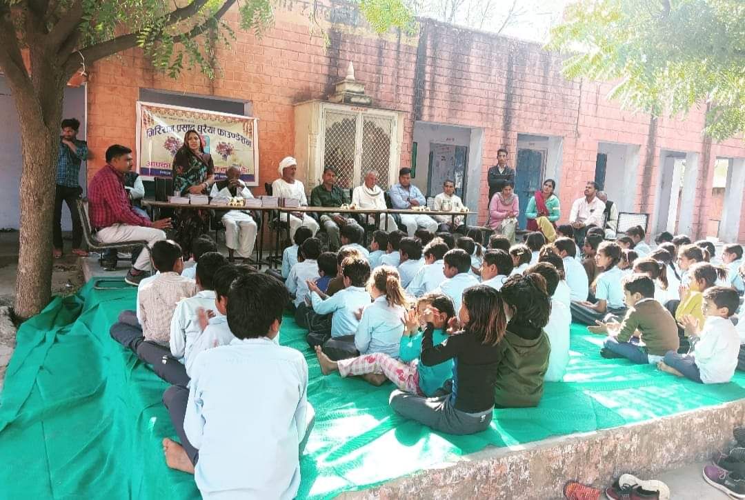 School children seated on a green mat in a courtyard listening to speakers at a table, beneath a foundation banner.