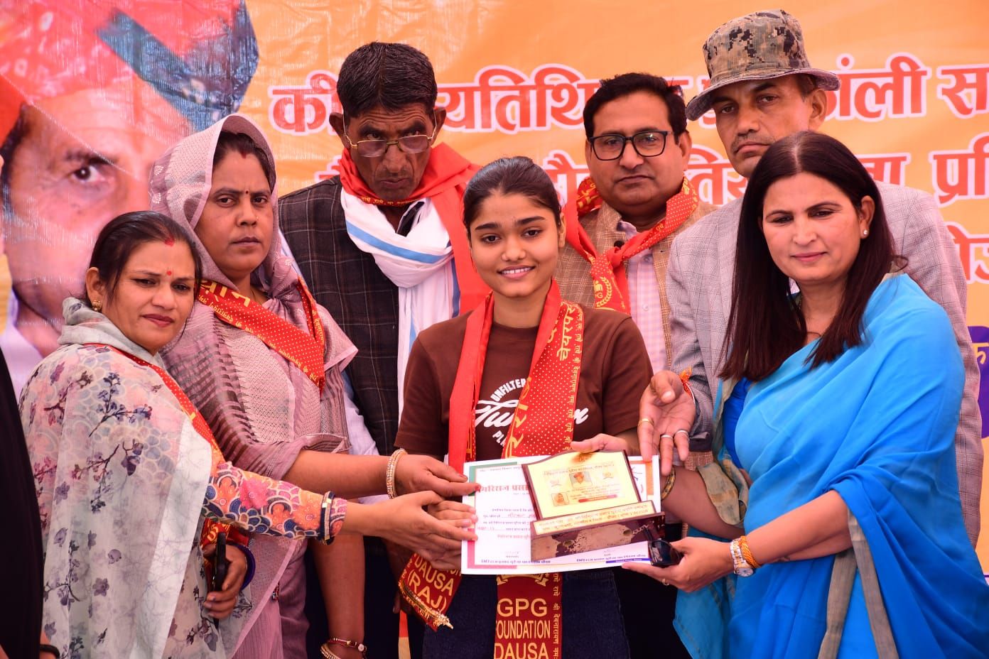 A student wearing a GPG Foundation Dausa sash receiving a certificate and a memento from guests at a ceremony.
