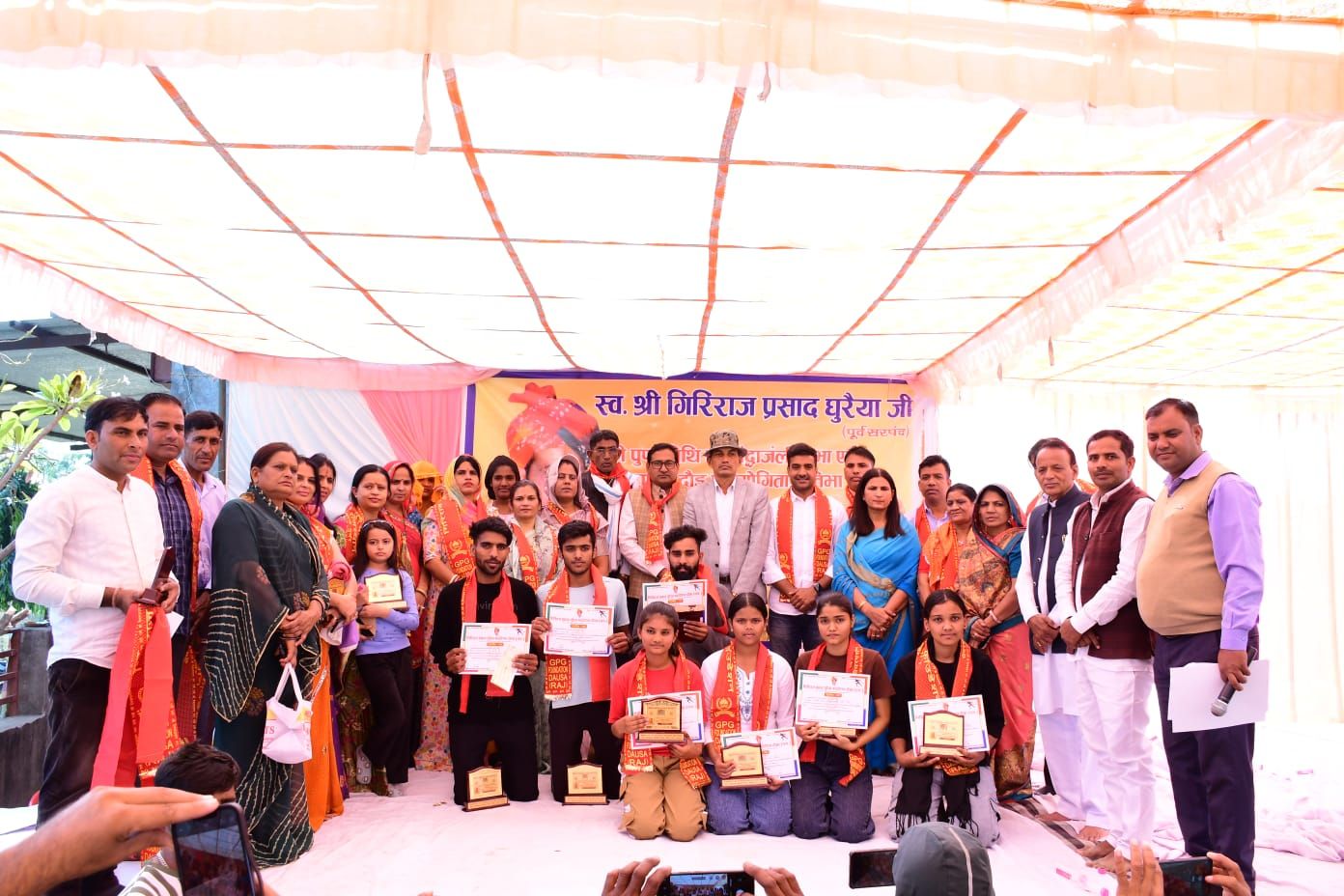 A large group photograph on a stage: students kneeling in front holding certificates and mementos, guests and organisers standing behind them.
